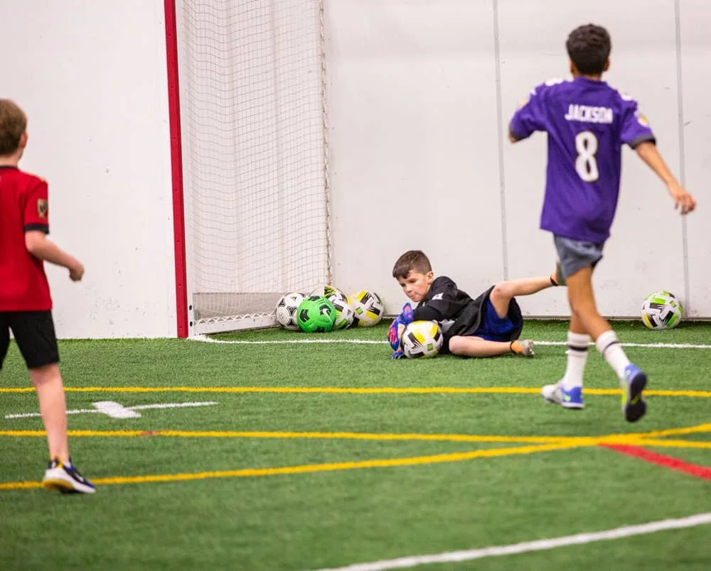 Young boys playing indoor soccer, with one kid in black goalie gear on the ground holding a soccer ball, other kids running around, and soccer balls near the goal.
