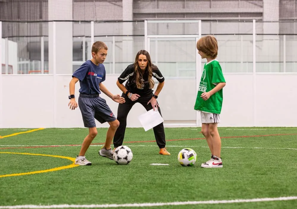 A soccer coach instructs two young boys during a practice inside an indoor soccer facility, with both boys standing on artificial turf and holding soccer balls.