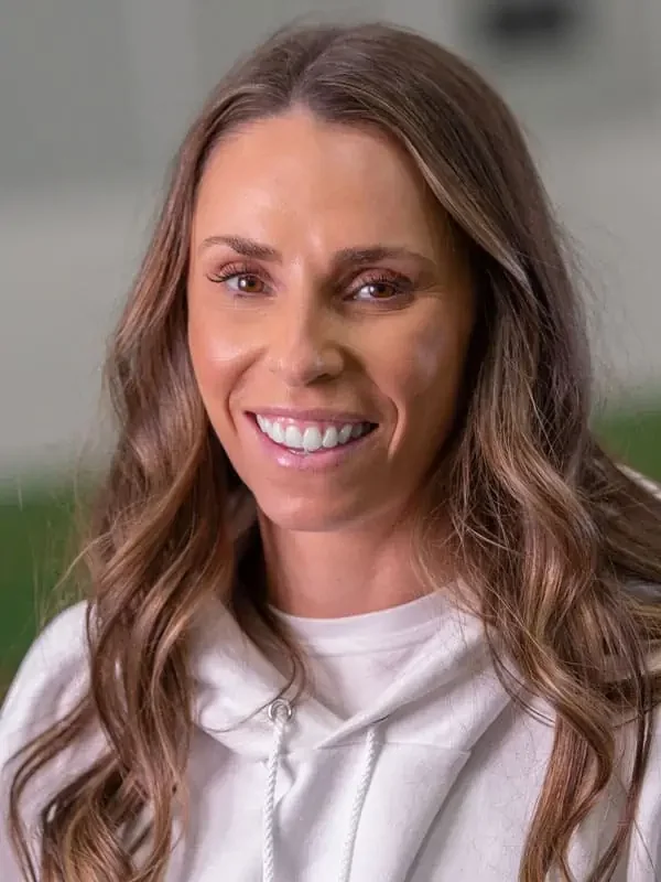 Close-up of a smiling woman with long brown wavy hair, wearing a white hoodie, indoors with blurred background.