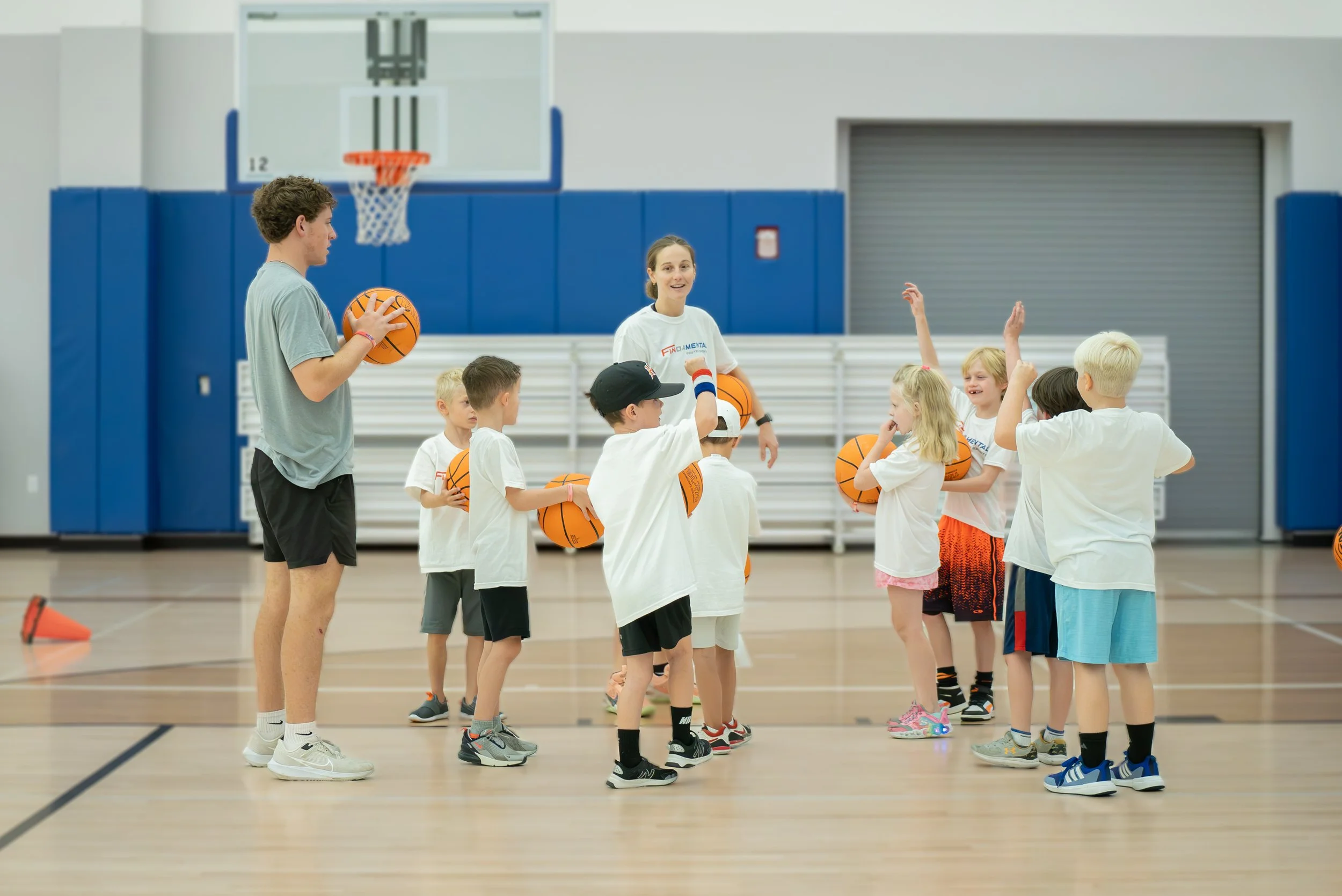 Children at a basketball practice in a gymnasium, with a coach giving instructions while holding a basketball. Some children are holding basketballs, with one raising their hand.