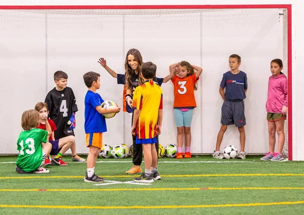 A group of children in athletic clothing and a woman coach on an indoor soccer field, some kids holding soccer balls, preparing for a practice or game.