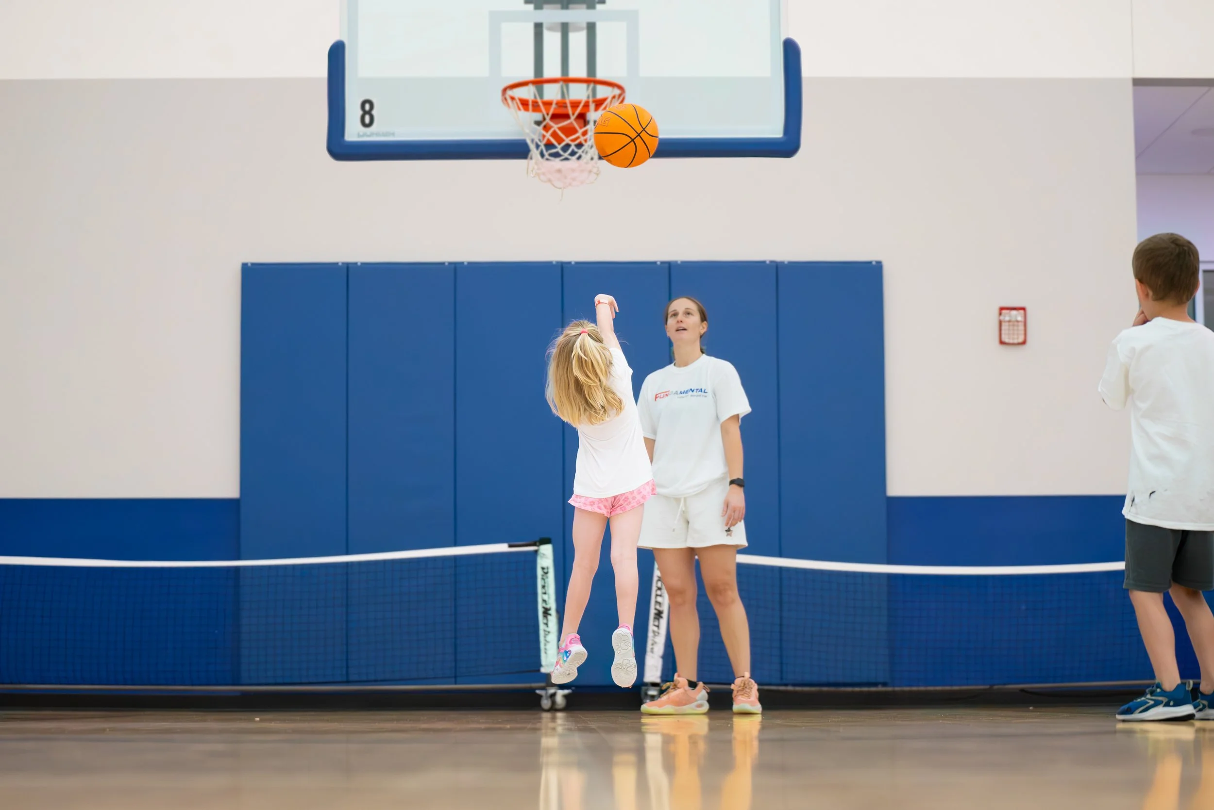 Children playing basketball in an indoor gym, with one girl jumping to shoot the ball and another girl standing nearby, while a boy observes in the background.
