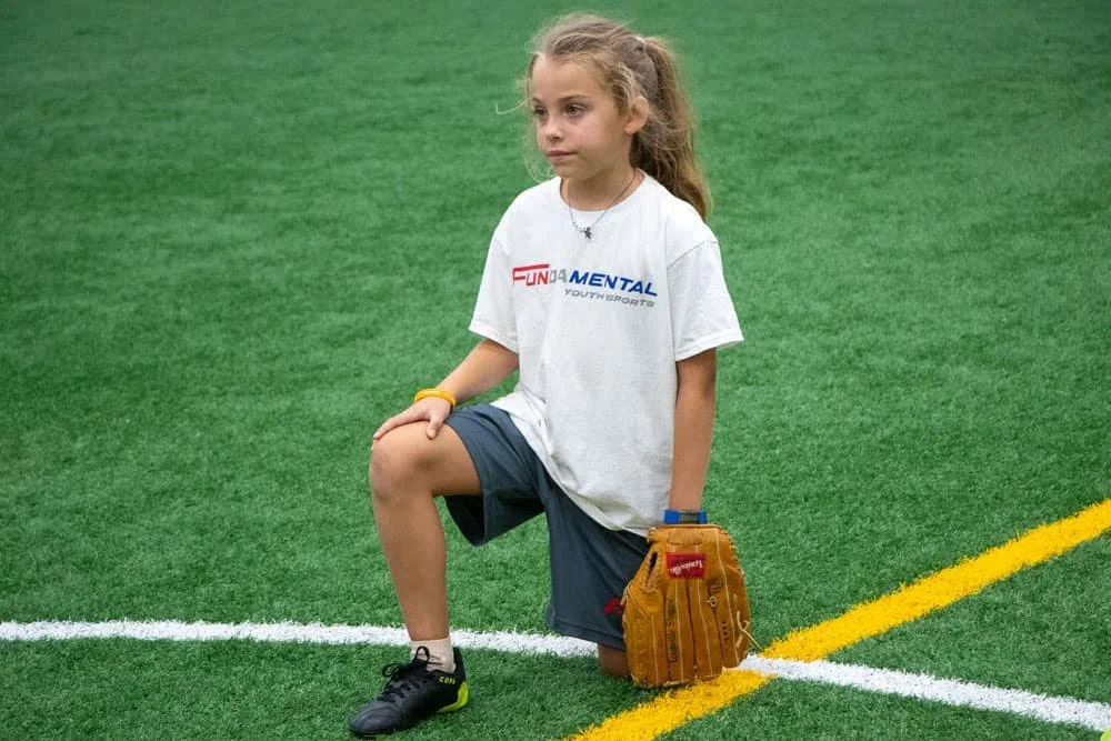 Young girl with a baseball glove kneeling on a green turf field.