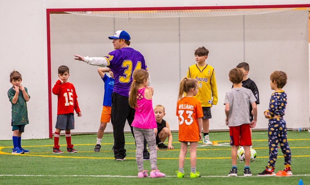 Children participating in a soccer practice indoors, standing on a field with a coach.