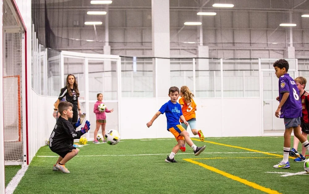 Children playing indoor soccer, with one child kicking the ball towards the goal and a goalkeeper preparing to catch it, others watching nearby.