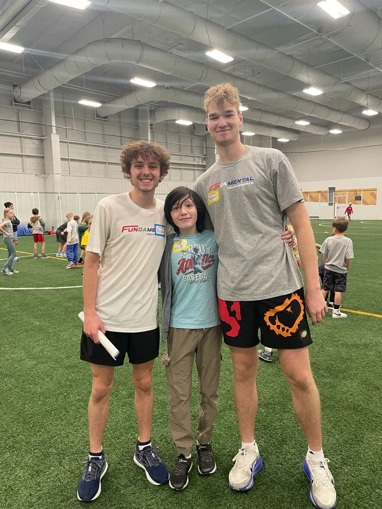 Three teenagers standing together inside an indoor sports facility on artificial turf, with other children playing in the background.
