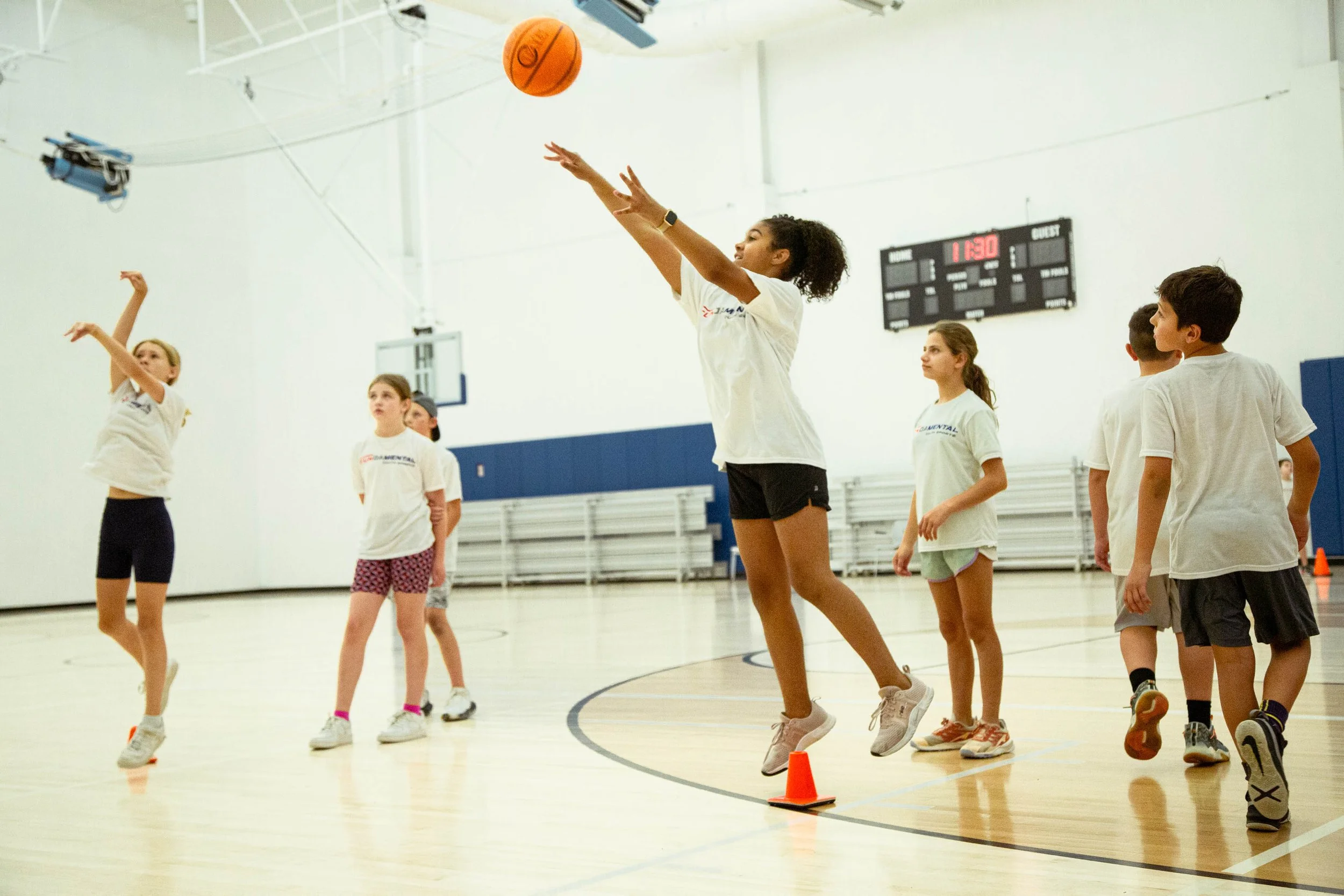 Children playing basketball in an indoor gym, with one girl shooting the basketball and others watching.