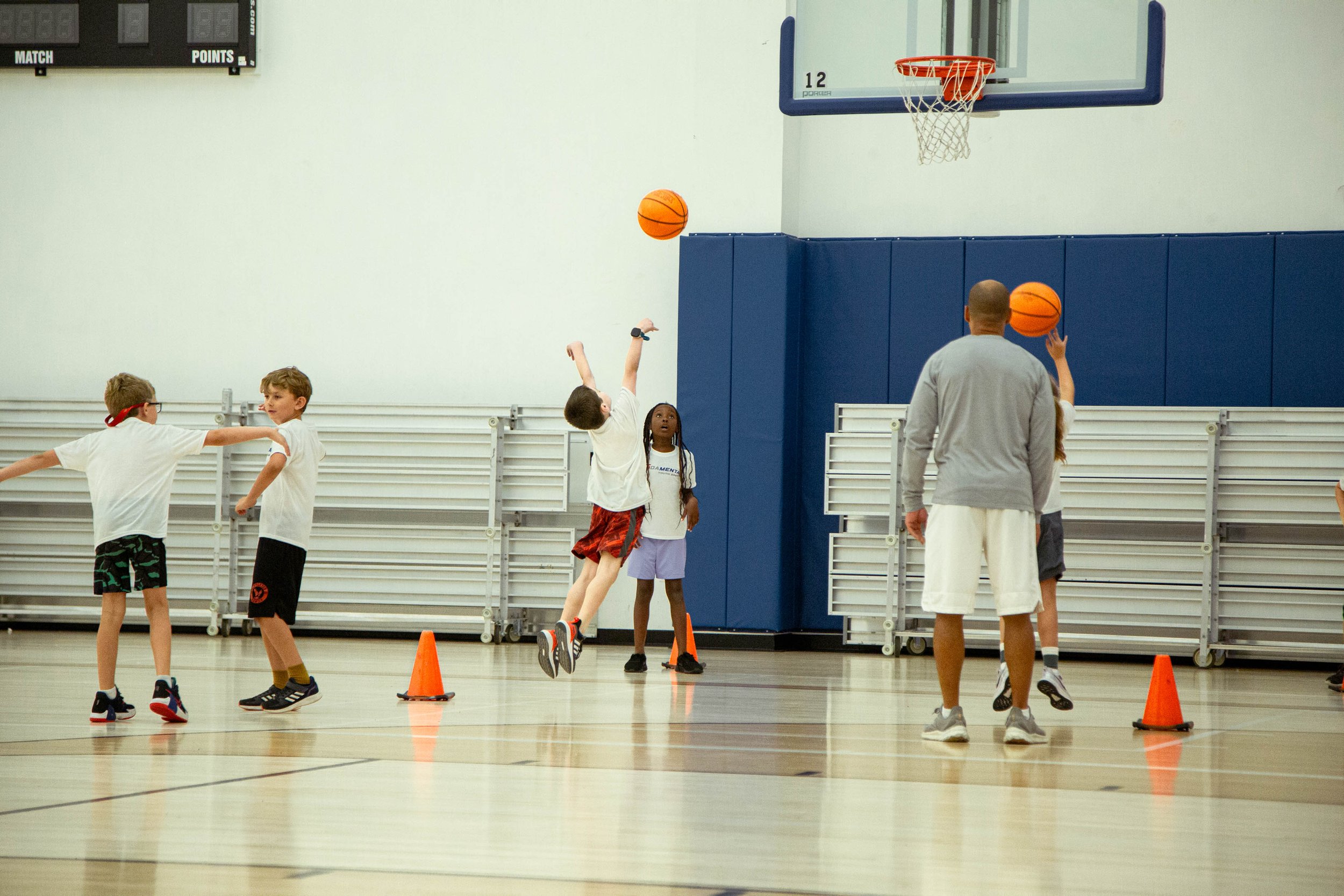 Children playing basketball in a gymnasium during a basketball practice or class, with an adult coach or instructor supervising.