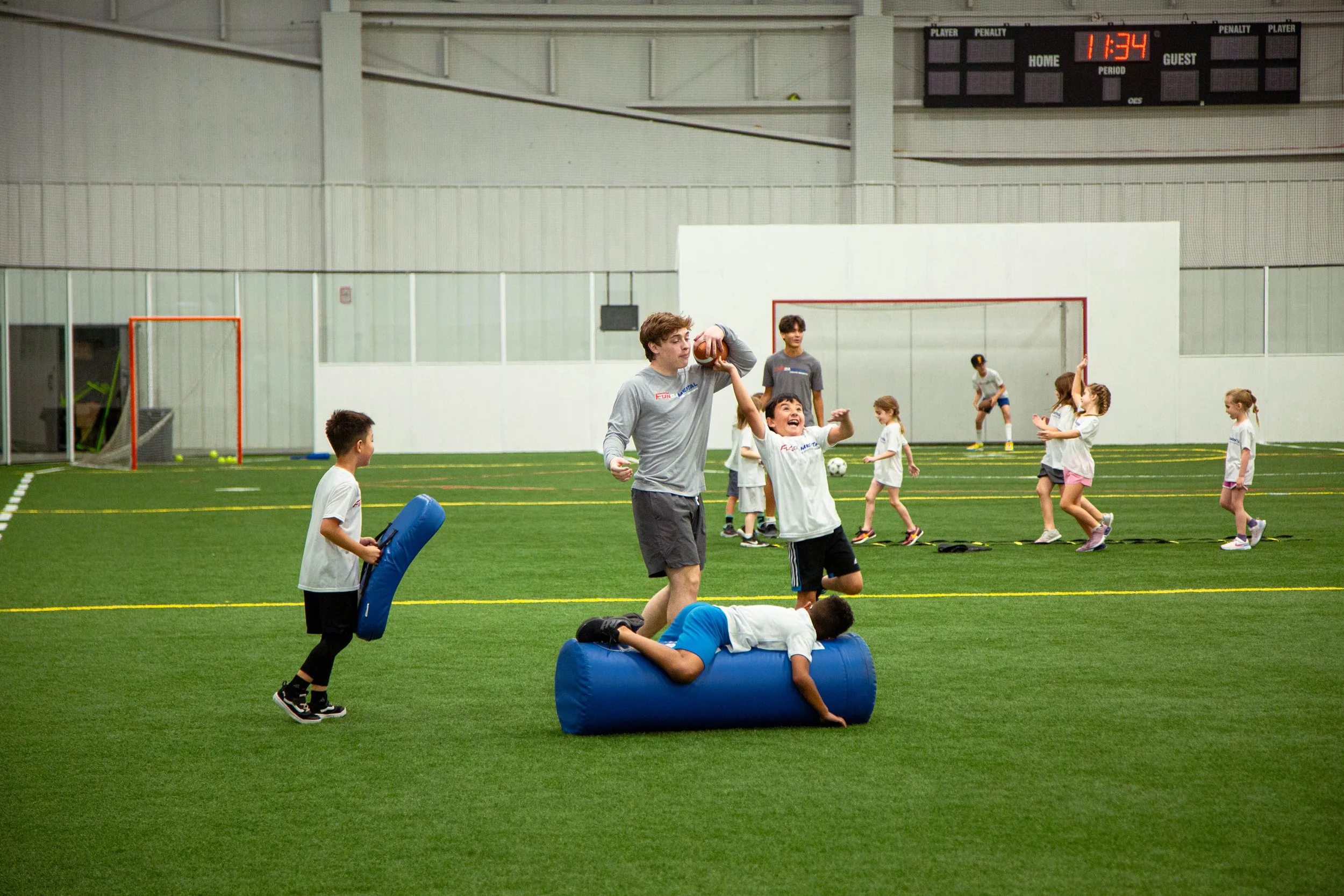 Children and teenagers participating in a football drill or training session inside an indoor sports facility. Some children are practicing running and jumping, while others are engaged in different activities.
