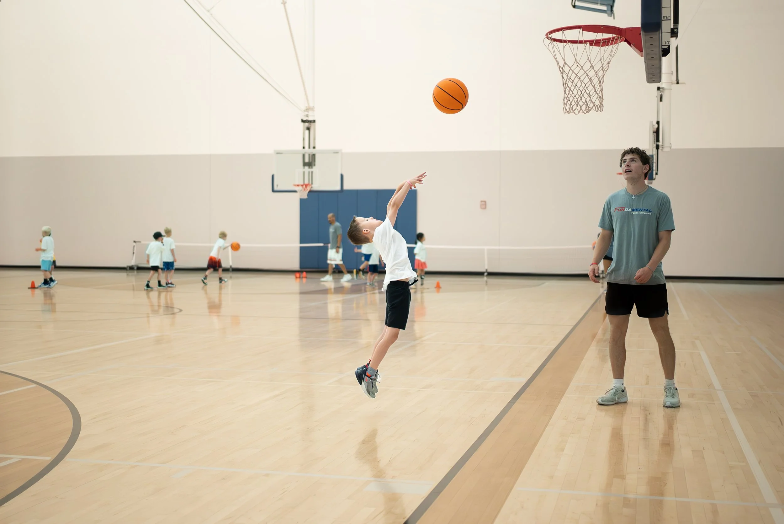 A group of children practicing basketball drills in an indoor gym, with one boy leaping to make a shot while a coach watches nearby.