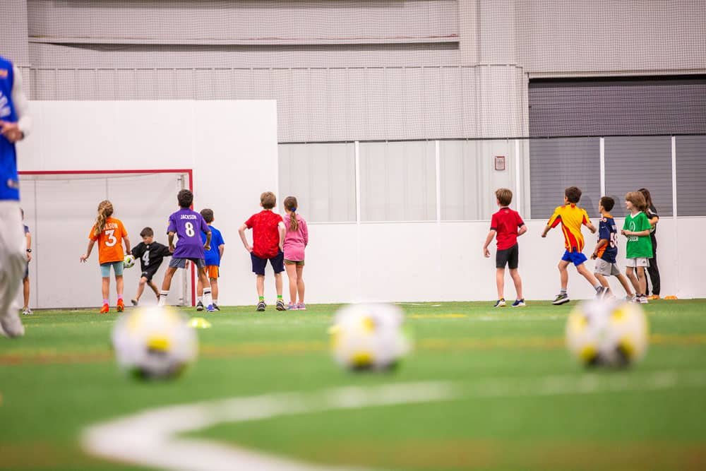 Children playing indoor soccer on a field with soccer balls in the foreground.