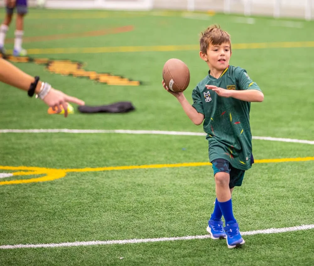 Young boy in green sports jersey running on an indoor football field while holding a football.