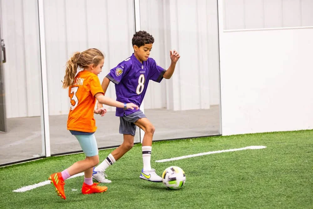 Two children playing soccer indoors. The boy is wearing a purple jersey, number 8, and the girl is in an orange jersey, number 3. They are competing for the ball on a green turf field.