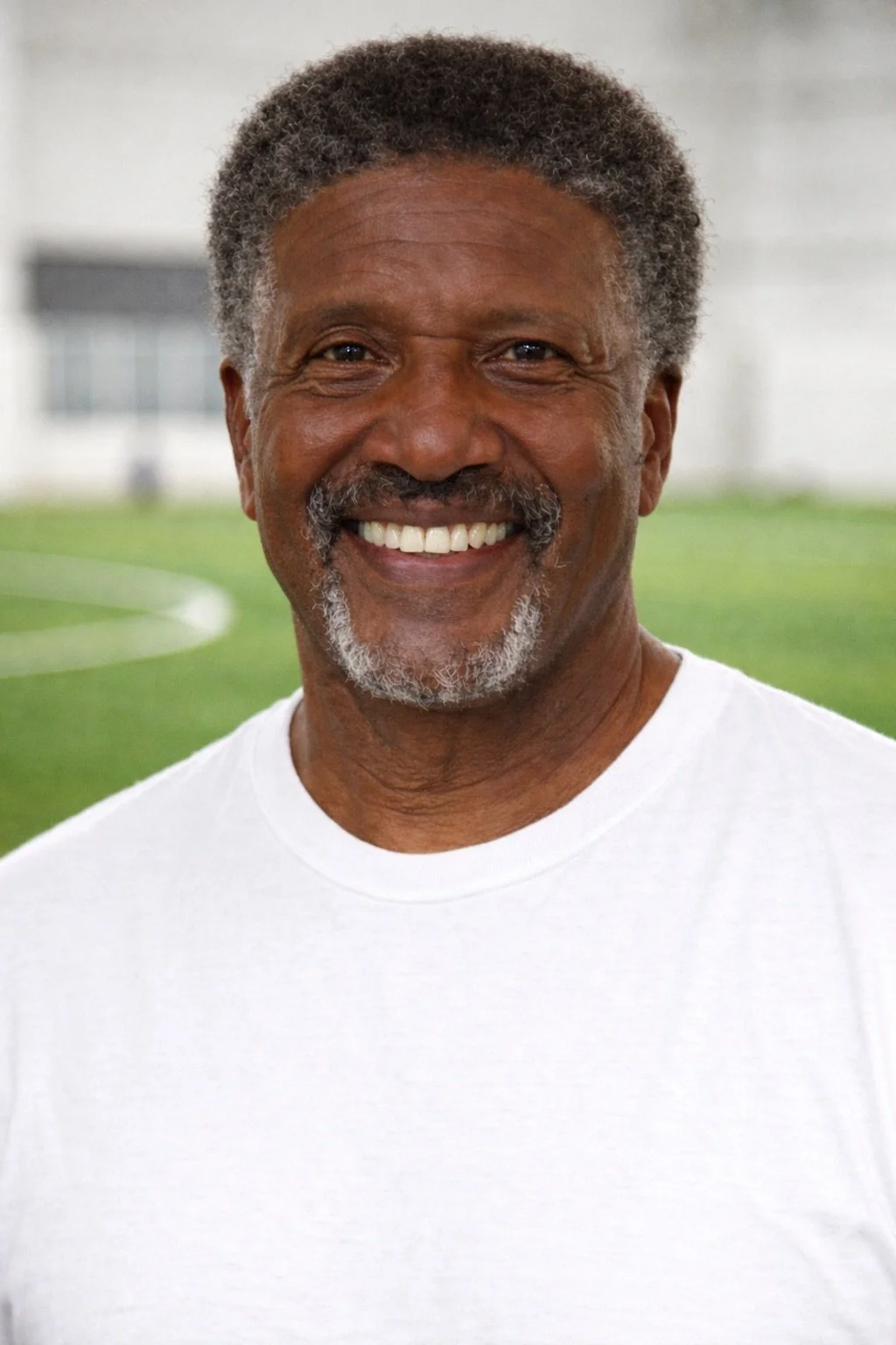Close-up of smiling middle-aged African American man with gray hair and beard, wearing a white t-shirt, standing on a soccer field.