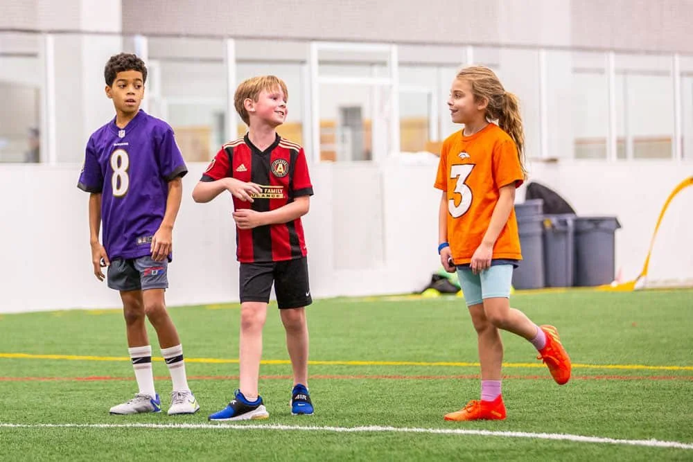 Three young children standing on an indoor sports field in sports jerseys, likely about to play a game.