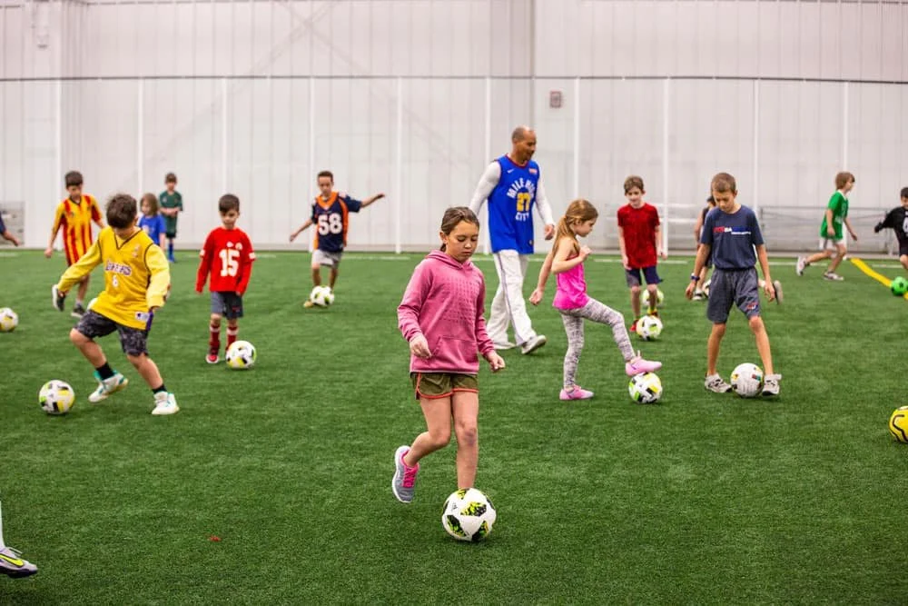 Children practicing soccer on an indoor field with an adult coach.