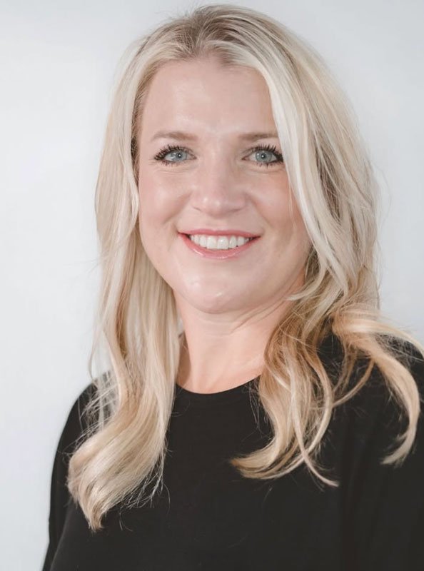 Portrait of a blonde woman with wavy hair smiling, wearing a black top against a light background.
