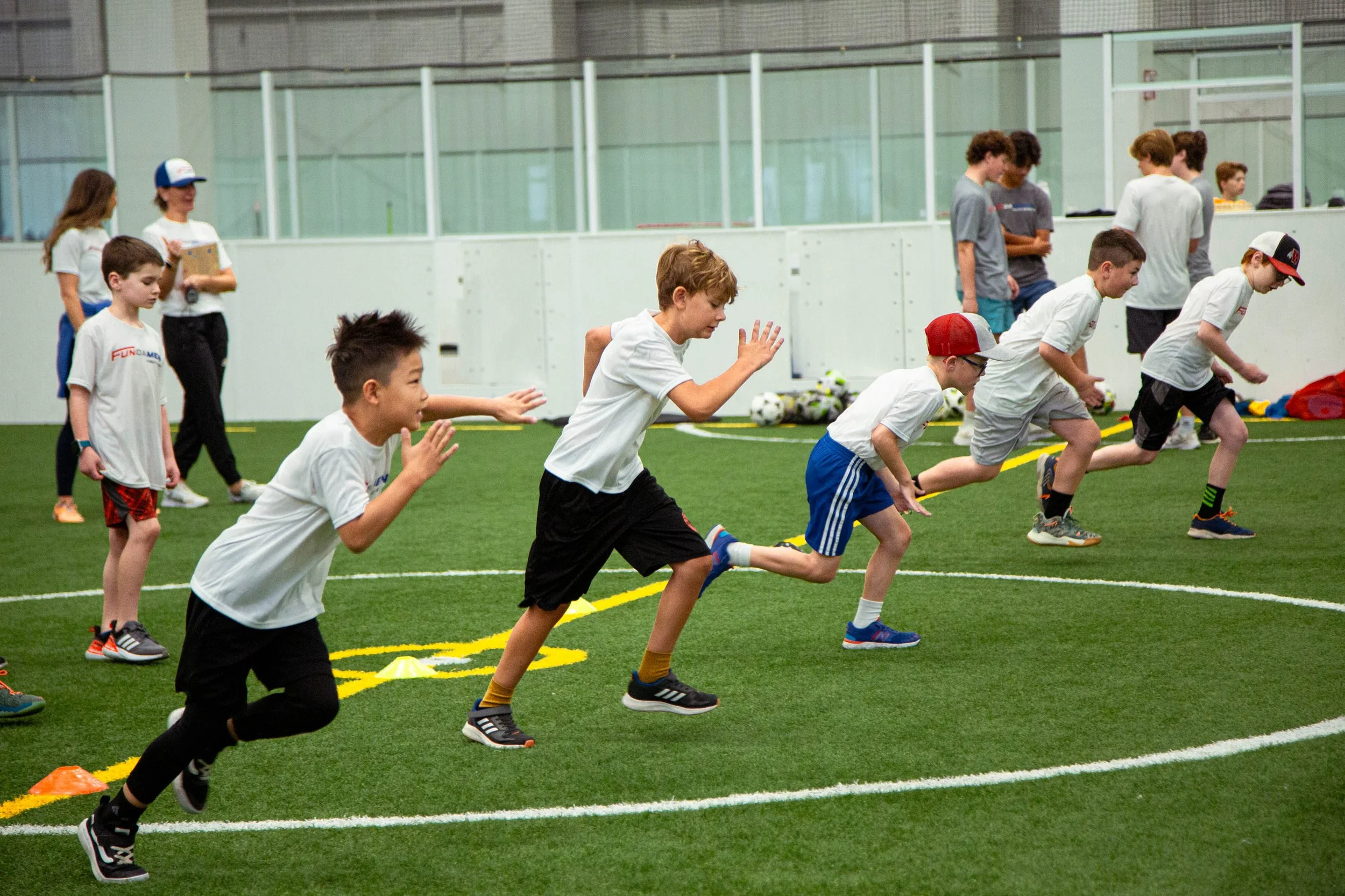 Children in sports attire racing at an indoor soccer practice or event, with coaches or spectators in the background.