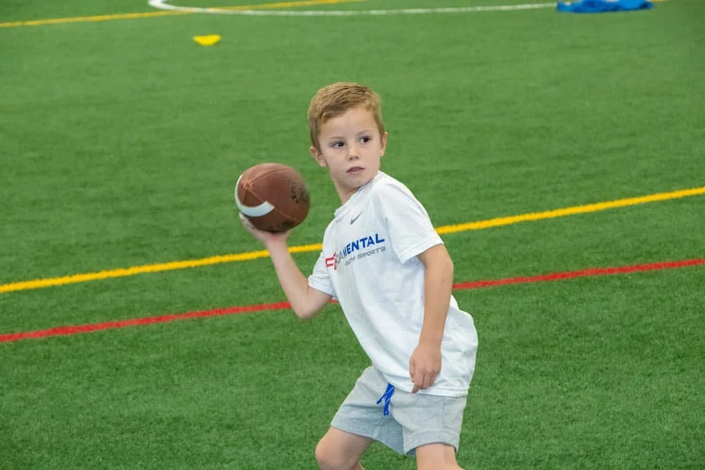 Young boy in white t-shirt and gray shorts holding a football on a green indoor sports field.
