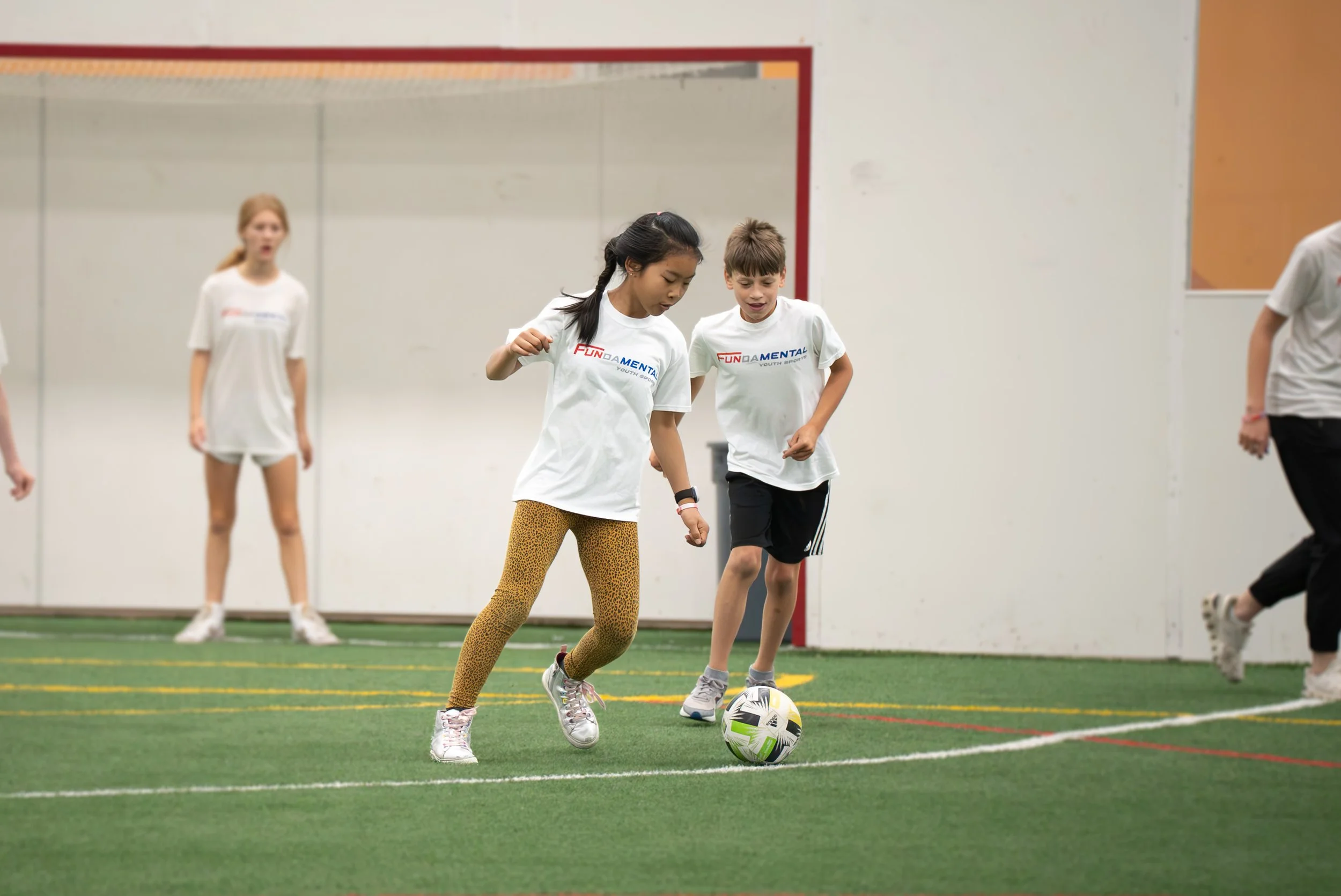 Children playing indoor soccer, two kids in white shirts and one girl in leopard print leggings, actively engaged in the game on a green turf
