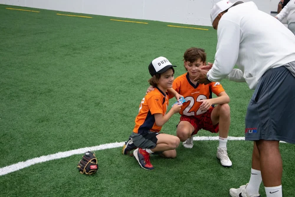 Two young boys in orange football uniforms sitting on the field, one with a baseball cap and one with a number 22 jersey, having a conversation with an adult man in athletic gear, with a football glove on the ground nearby.