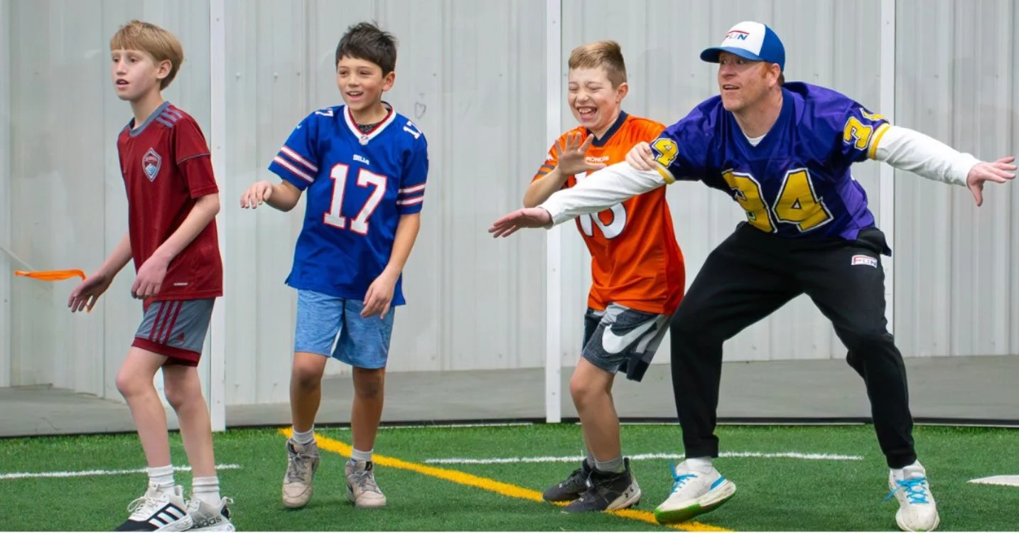 Four kids and one adult playing a game with a frisbee in a backyard with artificial turf and a metal fence.