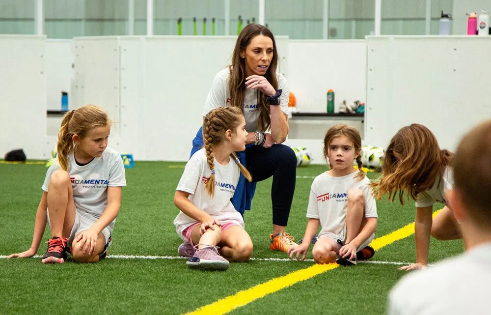 A coach crouching on the turf of an indoor soccer field, talking to a group of young girls seated and kneeling around her, during a youth soccer practice or game.