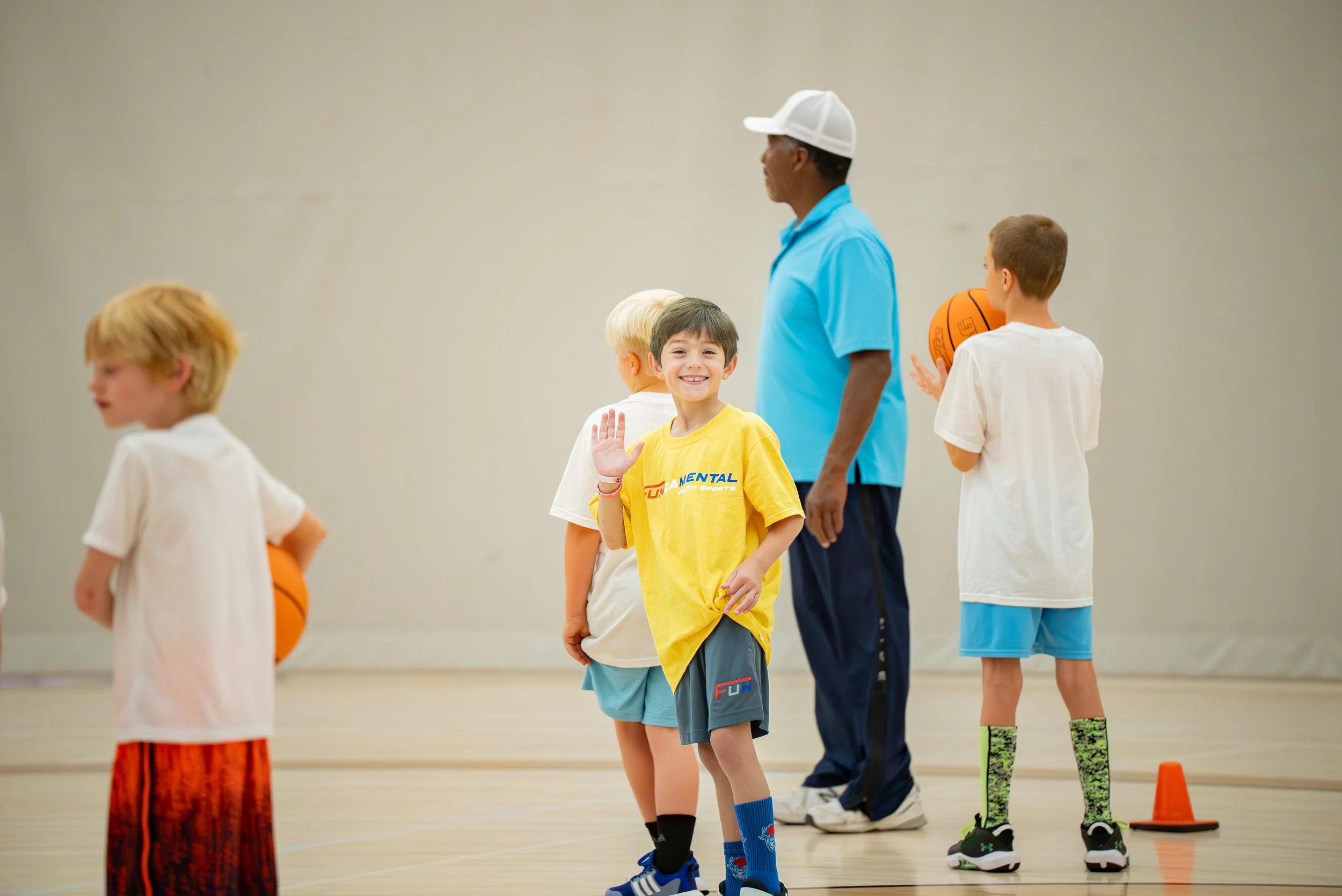 Children in a gym class learning basketball, with a coach supervising. The kids are dressed in sportswear, some holding basketballs, and orange cones are on the floor.