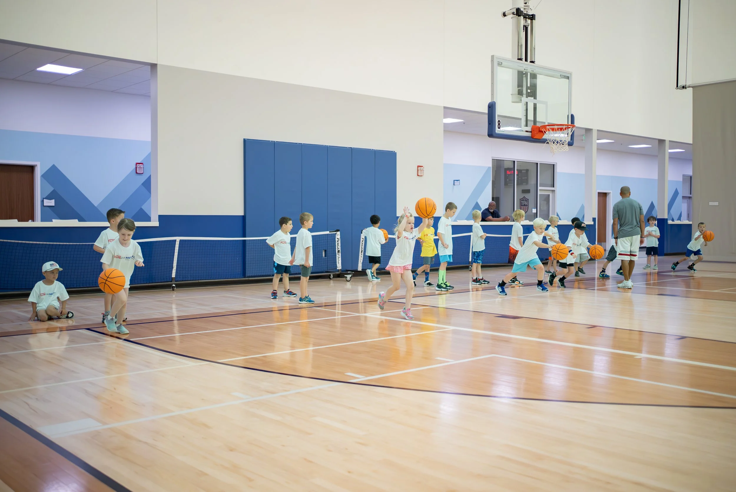 Children participating in a basketball training session in an indoor gym, some holding basketballs and others running or standing in line, guided by an adult coach.