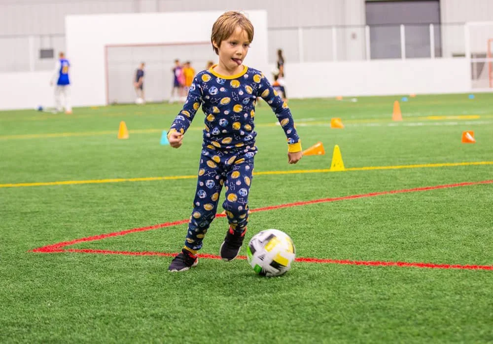 Young boy in pajamas playing soccer inside a sports facility with several orange and yellow cones on the artificial turf field.