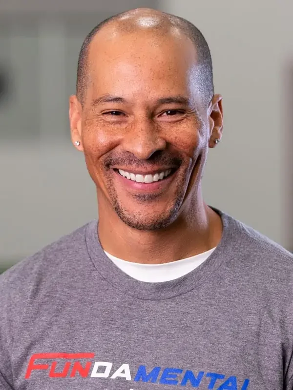 A smiling man with a shaved head and earrings, wearing a gray T-shirt with the words "FUNDA MENTAL" in colorful letters.