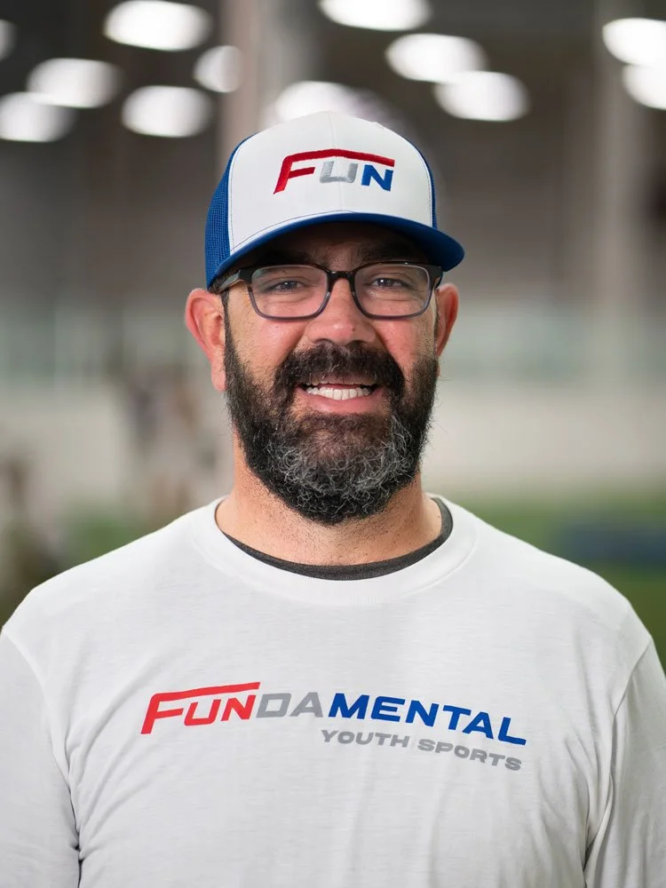 Smiling man with glasses and a beard, wearing a white T-shirt with 'FUNDA mental YOUTH SPORTS' logo, and a blue and white cap with 'FUN' logo, standing indoors with blurred background.