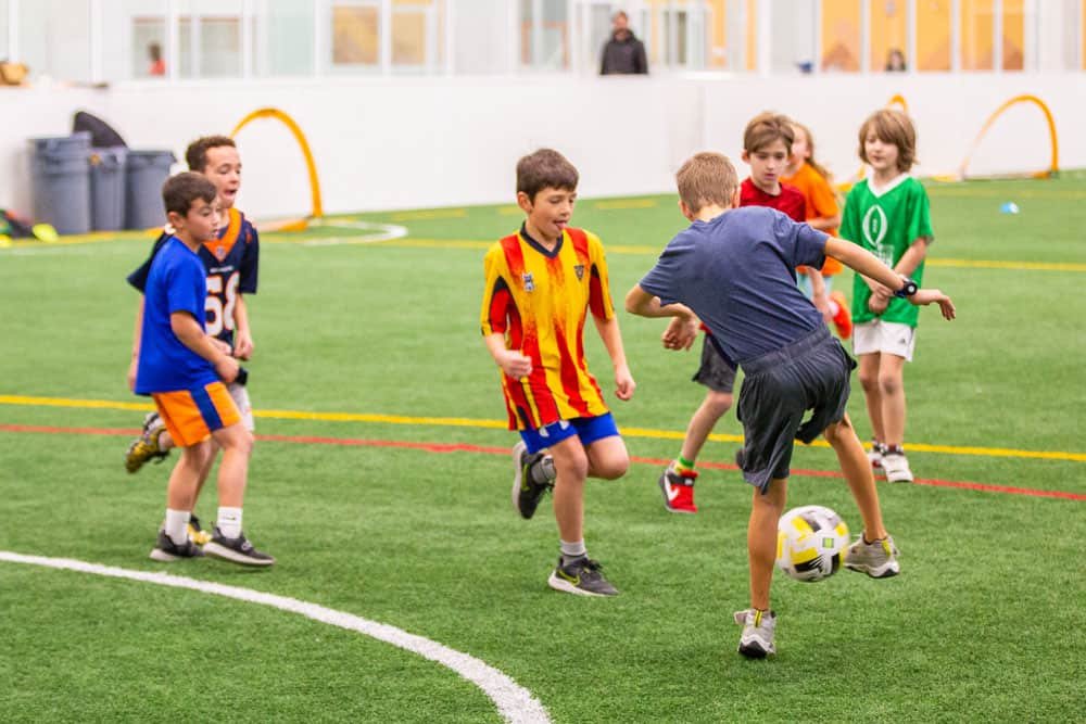 Kids playing indoor soccer on a field, with one child kicking the ball while others watch and cheer.