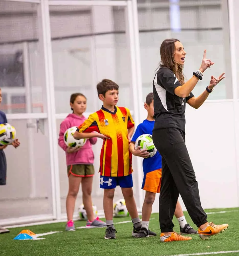 A female coach instructs young children during a soccer practice indoors, with children holding soccer balls and standing on a grassy field.
