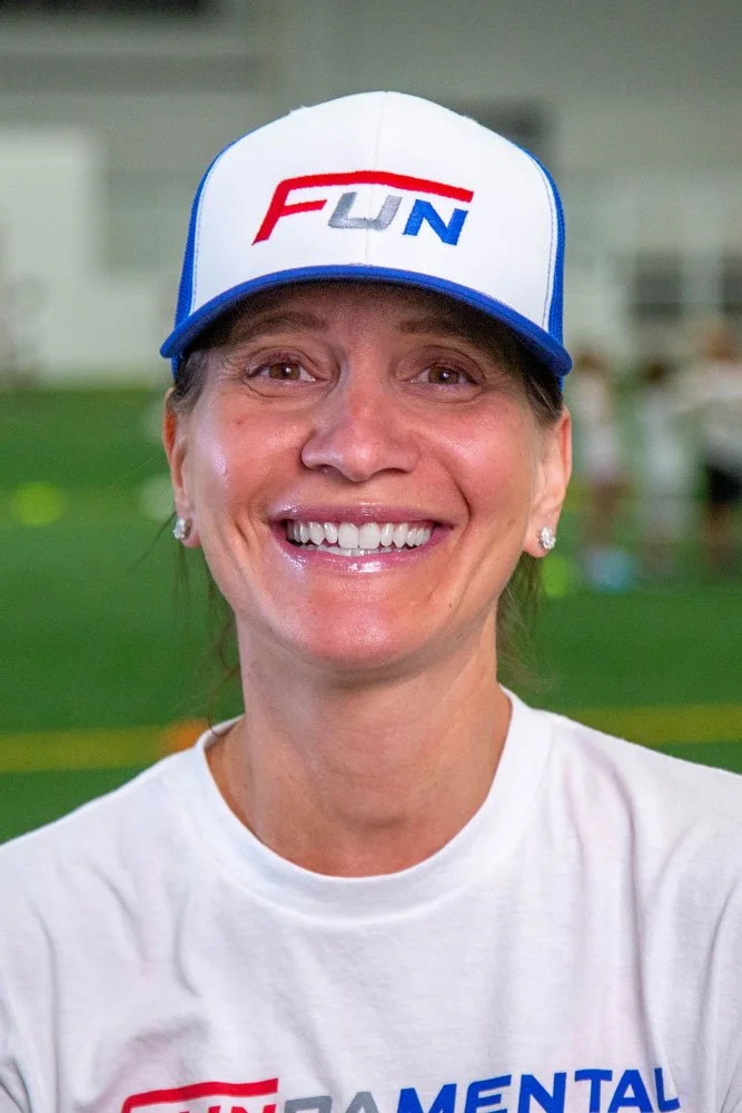 Close-up of a smiling woman wearing a white t-shirt and a white cap with 'FUN' logo, in an indoor sports facility with green field in background.