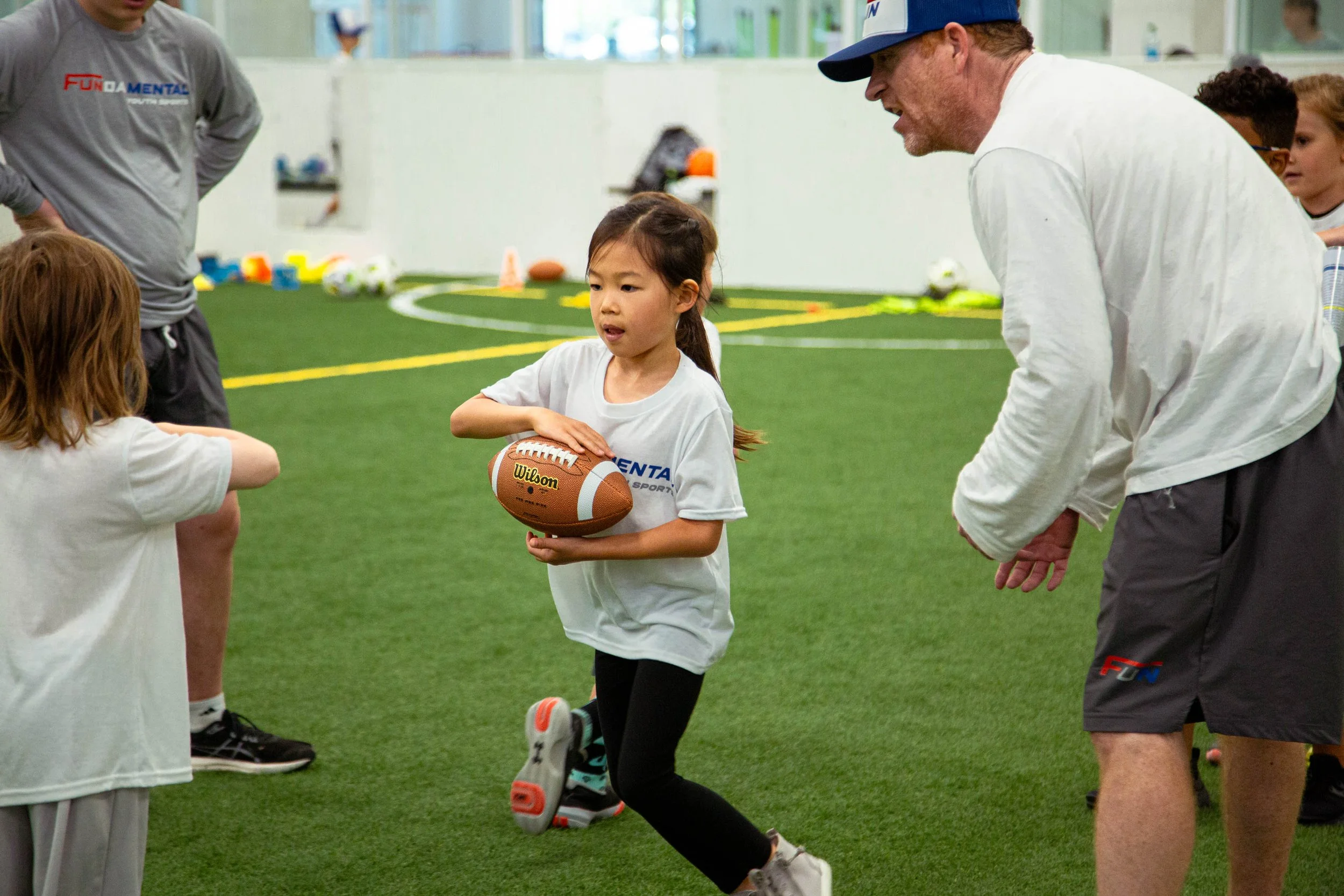 Young girl holding a football during a sports practice coach talking to her with other children around on an indoor field