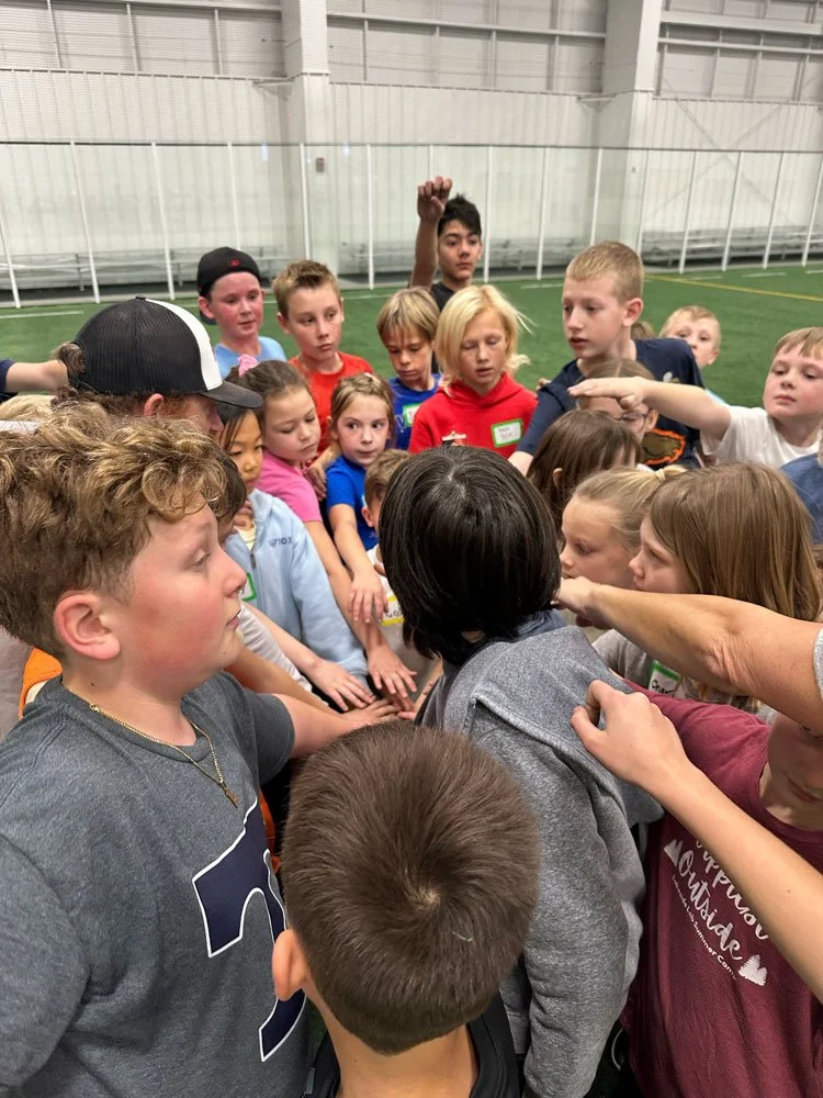 A group of children gathered together in an indoor sports facility, some with their hands on each other's shoulders, appearing to be engaged in a group activity or discussion, under the supervision of an adult.