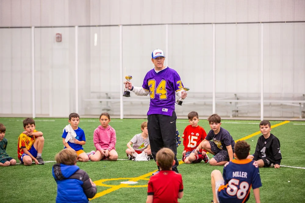 A coach in a purple jersey holding two trophies, standing in front of children seated on the indoor turf during a sports event or practice.