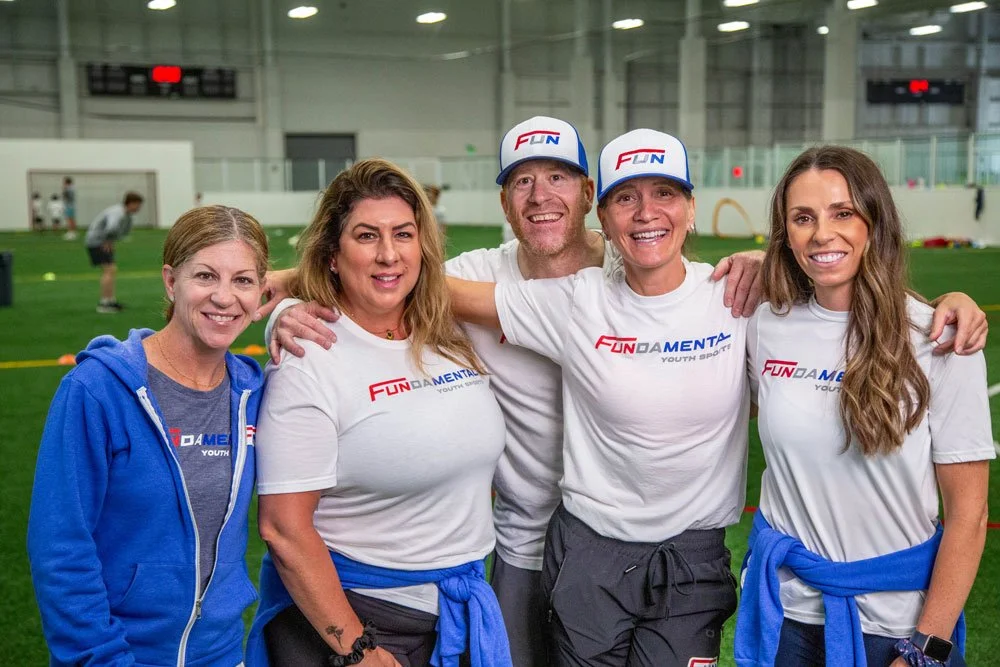 Group of five smiling adults at an indoor sports facility, wearing white T-shirts and caps with 'FUN DAMENTAL YOUTH SPORTS' logo, standing on a green field with children in the background.