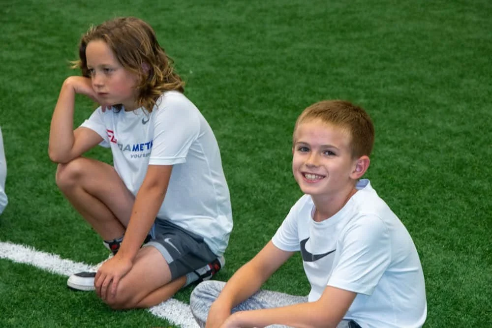 Two children sitting on artificial turf, one girl looking thoughtful and one boy smiling.
