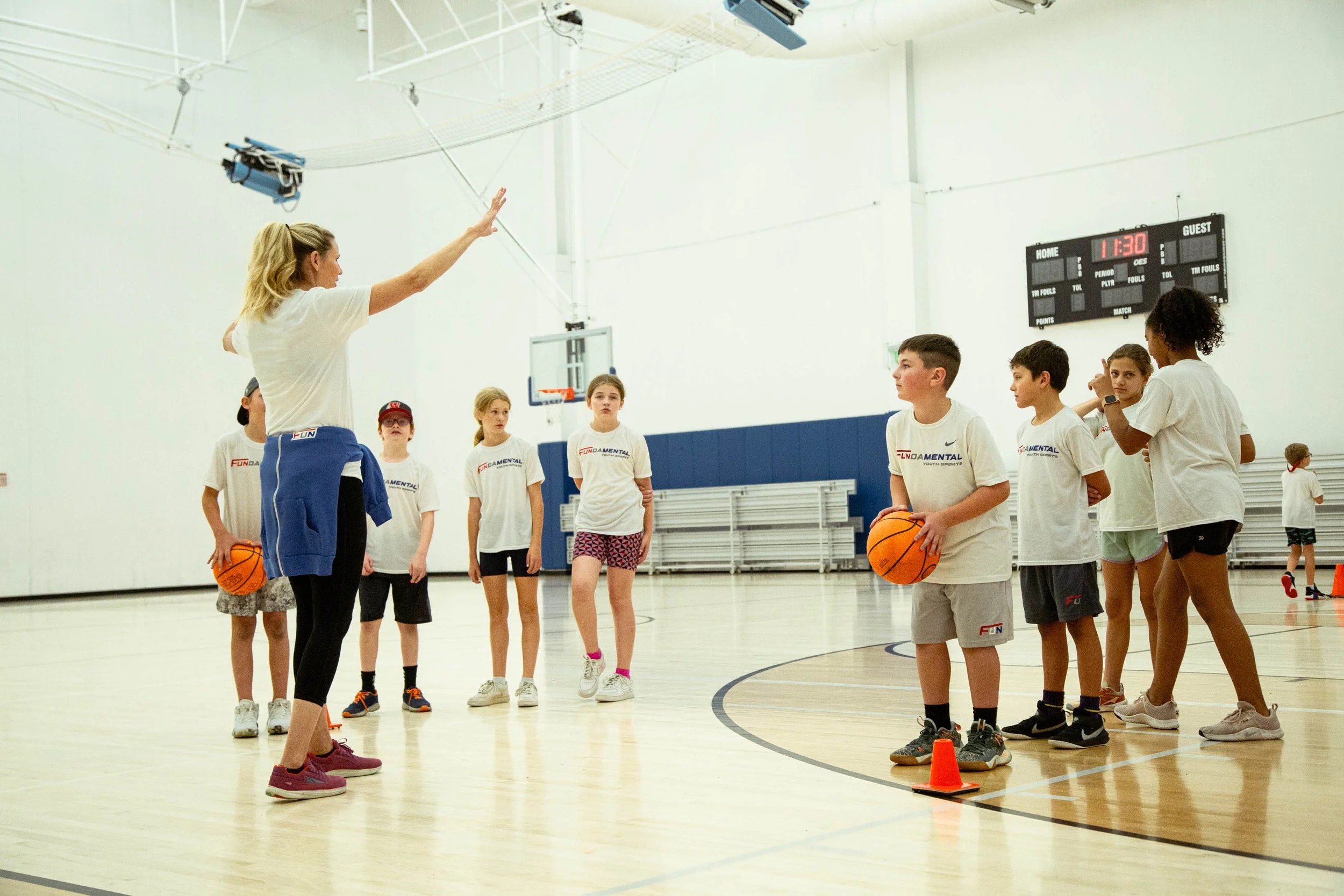 A basketball coach instructs young players during a practice session in an indoor gymnasium, with some kids holding basketballs and cones on the court.