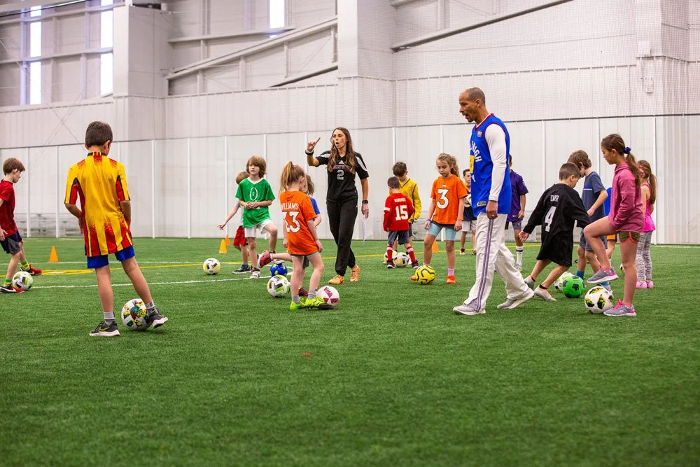 Children participating in a soccer training session on an indoor turf field, led by two coaches, one female and one male, with soccer balls and small orange cones.