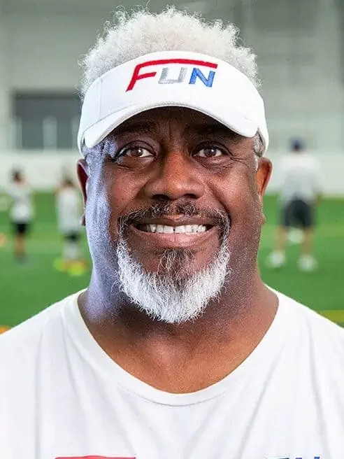 Smiling African American man with gray hair and beard wearing a white cap with 'FUN' logo and a white T-shirt, in an indoor sports facility with people in the background.