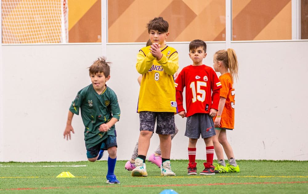 Kids dressed in sports jerseys standing on indoor turf field, preparing for a sports activity or practice.