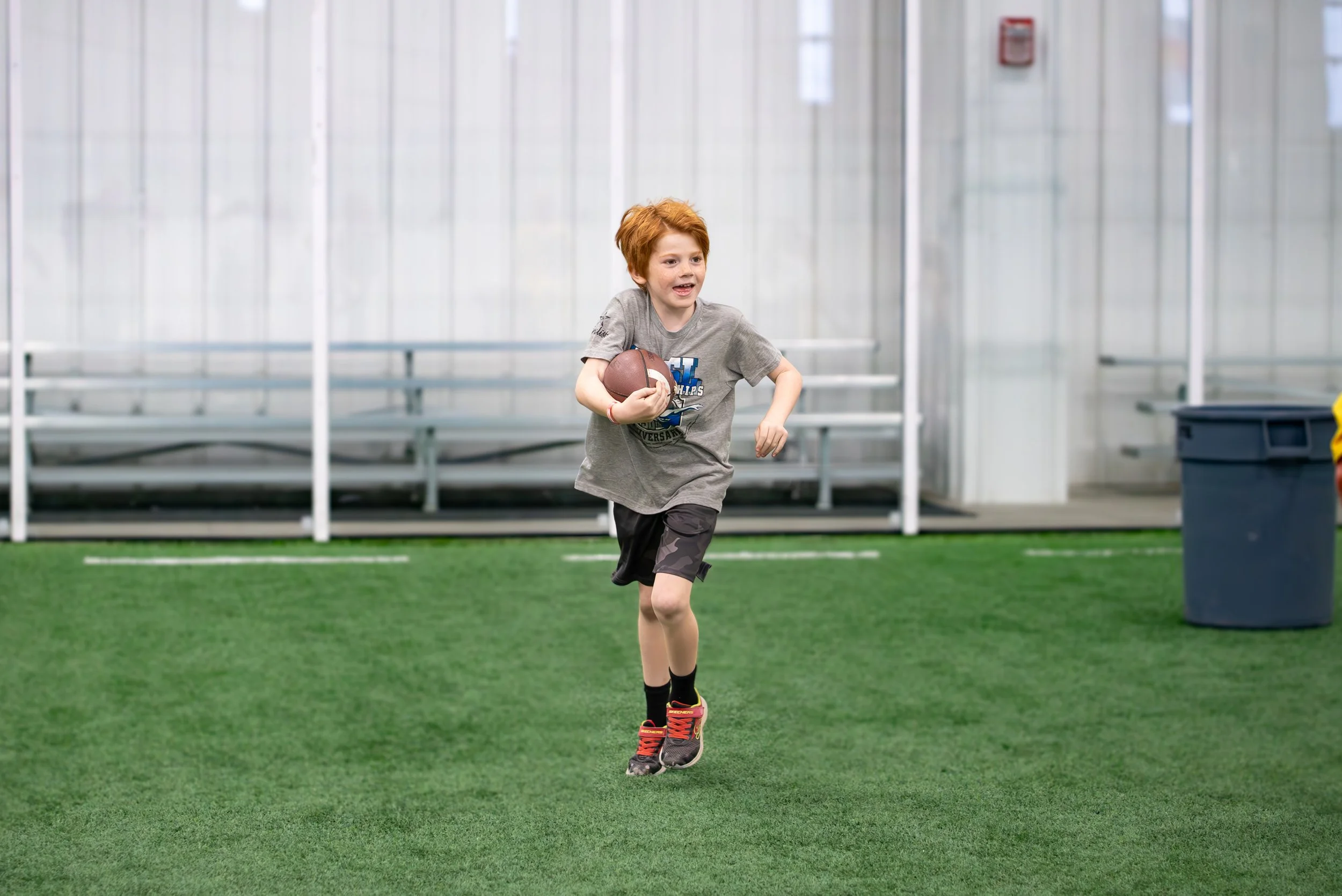 A young boy with red hair running on an indoor sports field while holding a football.