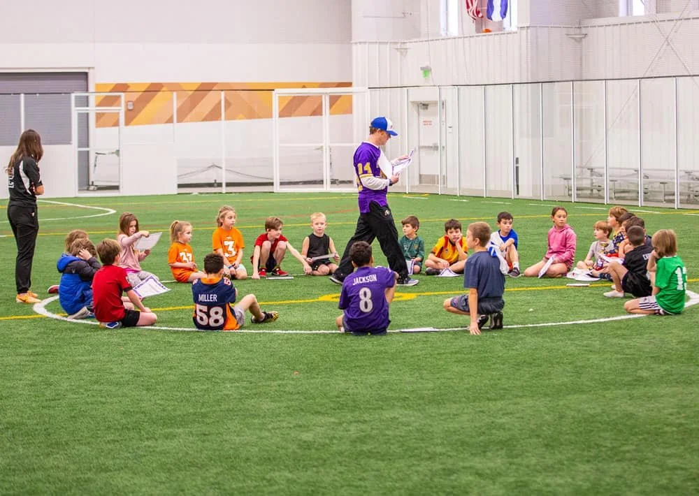 Group of young children sitting on artificial turf in a circle, listening to an adult coach holding a clipboard, inside an indoor sports facility.
