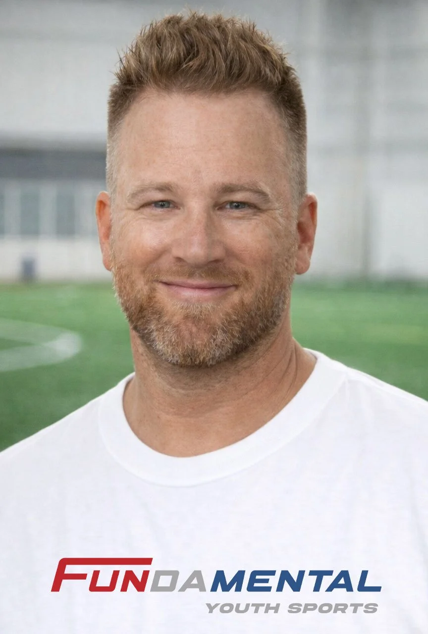 A smiling man with red hair and a beard, wearing a white t-shirt, standing outdoors on a sports field with a blurred background.