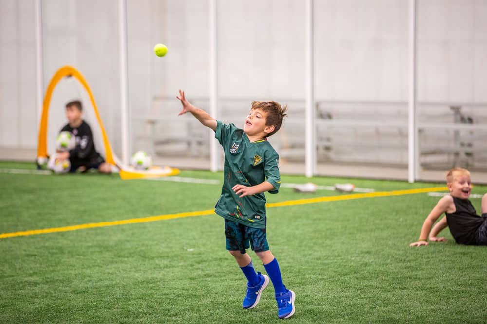 Young boy in soccer uniform catching a yellow tennis ball inside an indoor soccer facility.