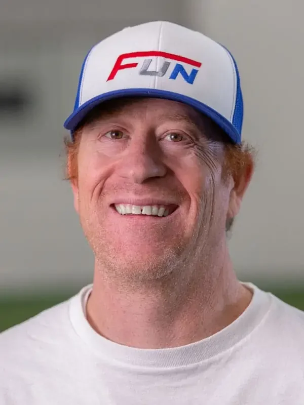 A man smiling, wearing a white t-shirt and a white and blue trucker hat with 'FUN' written on the front in red and blue letters.