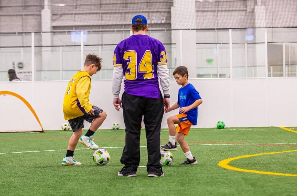 A coach teaches two young boys soccer skills on an indoor field.